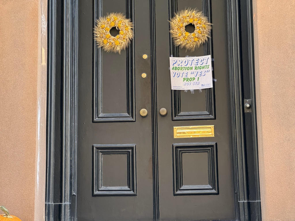 Una puerta negra de doble hoja con detalles clásicos está decorada con dos coronas de flores amarillas secas. Un cartel en el centro aboga por "PROTECT ABORTION RIGHTS" y solicita votar "YES" en la Proposición 1, con la fecha "NOV 5TH". Una calabaza es visible en la esquina inferior izquierda. (Foto: Ismario Rodríguez / Periodismo de Barrio).