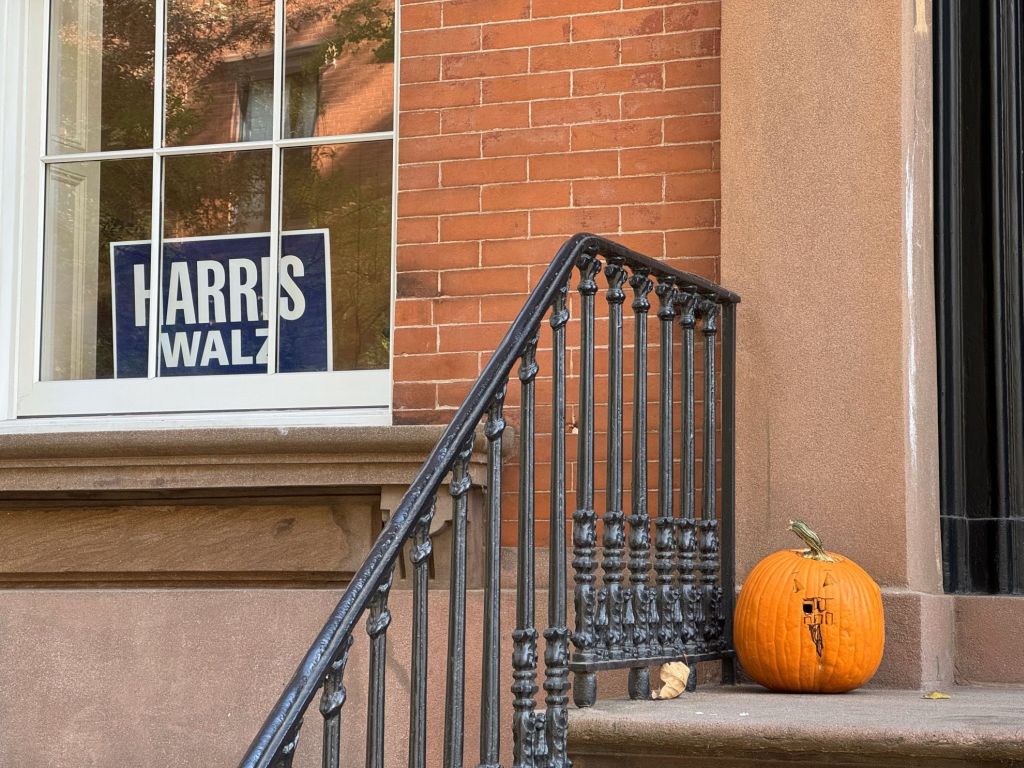 Una calabaza tallada con un diseño minimalista descansa sobre un escalón junto a una barandilla negra de hierro forjado. En el fondo, una ventana muestra un cartel electoral azul con los nombres "HARRIS WALZ" en blanco, sobre una pared de ladrillos rojizos. (Foto: Ismario Rodríguez / Periodismo de Barrio).