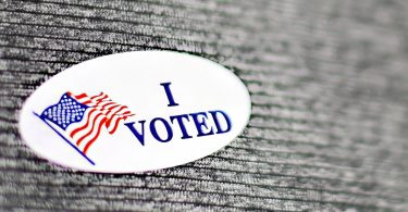 Una pegatina ovalada blanca con las palabras "I VOTED" en letras azules y una bandera estadounidense ondeante en el lado izquierdo, sobre un fondo texturizado gris oscuro. Foto: Imagen de Pixabay.