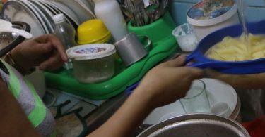 Una mujer escurre pastas en un colador azul sobre un fregadero lleno de utensilios y recipientes plásticos.