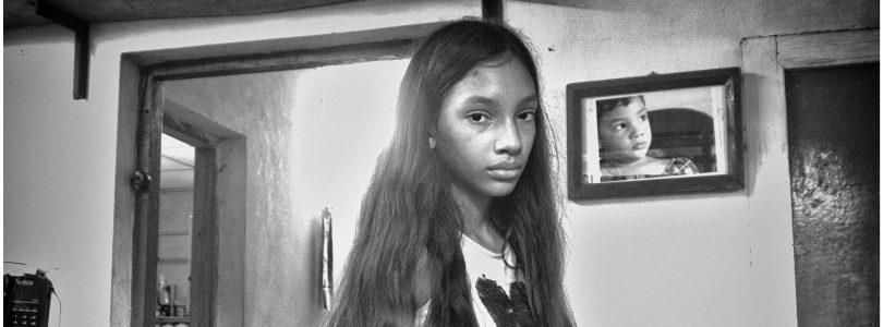 Niña con expresión seria en el interior de una casa modesta, con un retrato infantil en la pared.