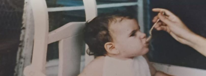 Bebé sentada en una silla alta de madera mientras una mano adulta le da de comer con una cucharita.