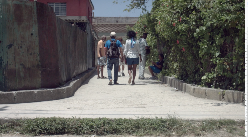 Zaima y Tomás caminan juntos por un pasillo estrecho en su barrio, rodeados de otras personas, vegetación y estructuras deterioradas.