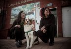 Geraidy Brito Montes de Oca y Yaqui Saiz, junto a su perro, sentadas frente al mural de la Casa de Títeres Cubana Argentina en Junín. Detrás, se ve la fachada del espacio cultural con puertas blancas y paredes de ladrillo rojo. Foto: Ella Fernández.