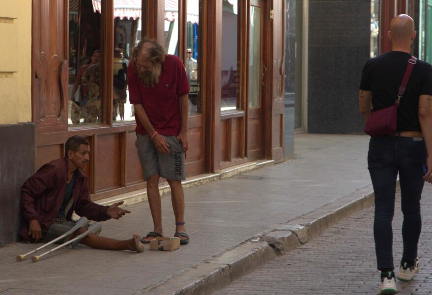 Dos hombres en situación de calle en un portal; uno sentado con muletas, otro de pie; transeúnte al fondo.