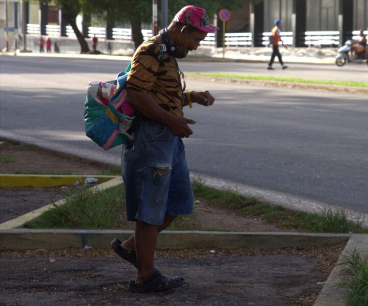 Hombre con gorra rosa, audífonos al cuello y mochila remendada cruza una avenida.