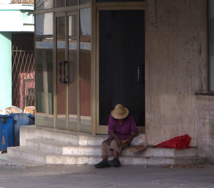 Persona con sombrero y camisa morada sentada en los escalones de un portal, junto a bolsa y sombrilla roja.