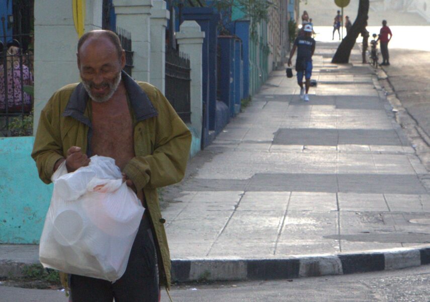 Hombre mayor camina por la acera con una bolsa plástica blanca y chaqueta abierta.
