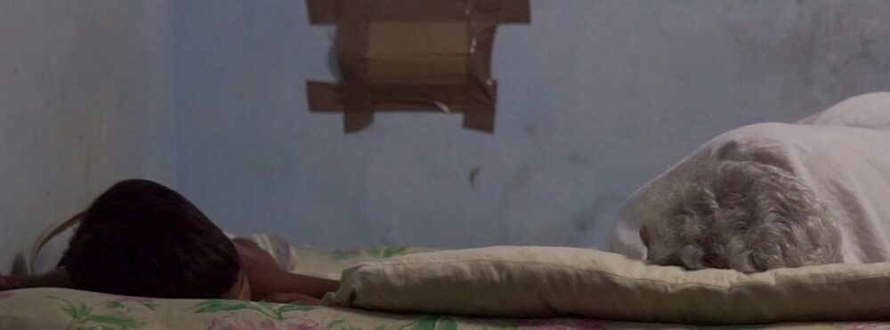 Hacinamiento en Santiago de Cuba. Dos personas (un niño y una adulta mayor) duermen en una cama estrecha en un cuarto pequeño y oscuro, simbolizando el agotamiento de una madre soltera en Cuba.