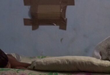 Hacinamiento en Santiago de Cuba. Dos personas (un niño y una adulta mayor) duermen en una cama estrecha en un cuarto pequeño y oscuro, simbolizando el agotamiento de una madre soltera en Cuba.
