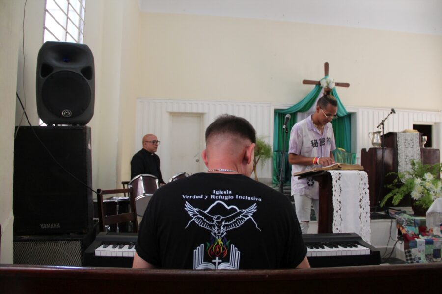 Hombre adulto de espalda al espectador toca teclado musical durante acto religioso en interior de iglesia. De fondo se ve sacerdote, Cruz de madera con tela verde, plantas decorativas y equipamiento de audio