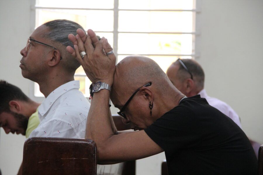 Primer plano de un hombre adulto con lentes de marco oscuro, con sus manos juntas cerca del rostro en actitud de oración o reflexión profunda, luz natural que entra por ventana de fondo