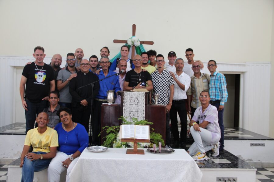 Grupo diverso de aproximadamente de personas de distintas edades y géneros posados frente a un altar con cruz de madera, biblia abierta y flores, en interior de templo blanco