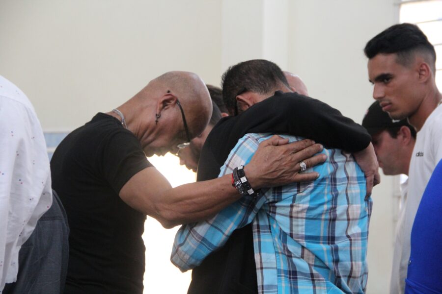 Grupo de hombres rodean a una persona en el centro, colocando sus manos sobre su espalda y hombro en gesto de apoyo y consuelo durante un acto religioso