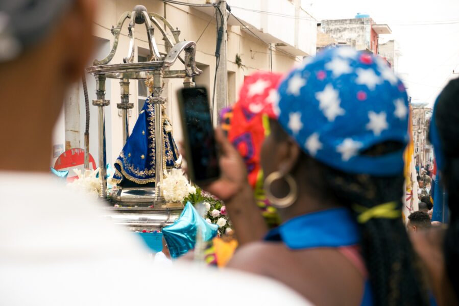 Virgen de Regla siendo fotografiada por una mujer cubana que lleva una gorra alegórica a la bandera de Estados Unidos. Foto: Neus Pechero.