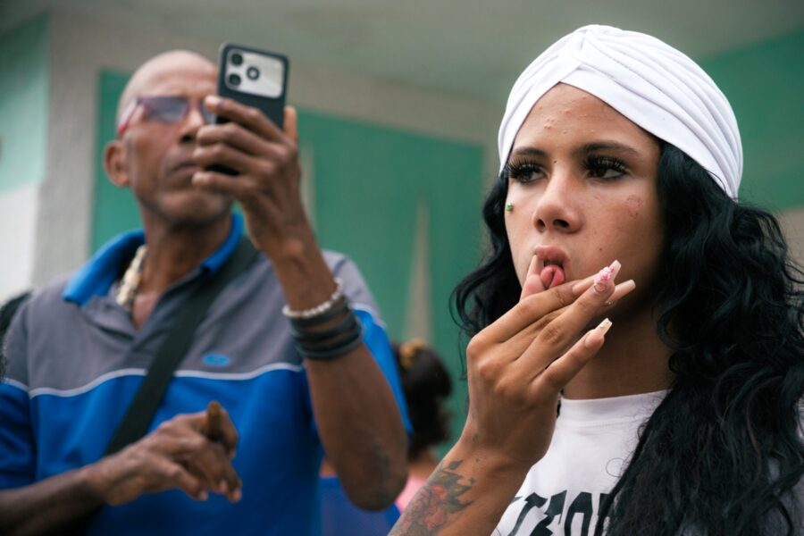 Joven cubana observa la procesión de la Virgen de Regla desde la acera mientras otra persona documenta lo que sucede. Foto: Neus Pechero.